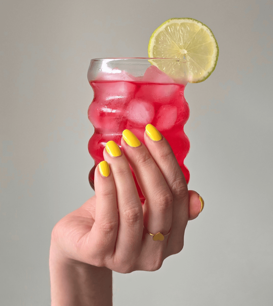 A hand wearing bright yellow indie nail polish holds a summer drink with a slice of lime in it