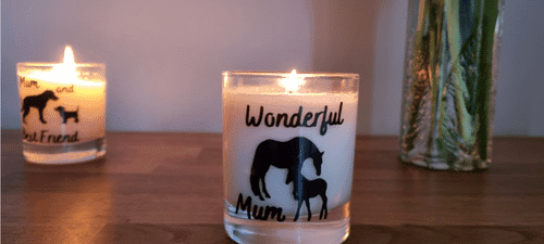 A glass container candle with white wax. The container has a horseand foal design in black and the words wonderful mum, the candle is lit.