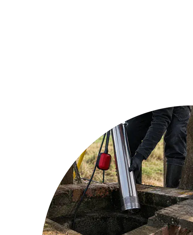 Technician lowering a borehole submersible pump into a rural brick well, with connected hoses and cables in an outdoor installation setting.