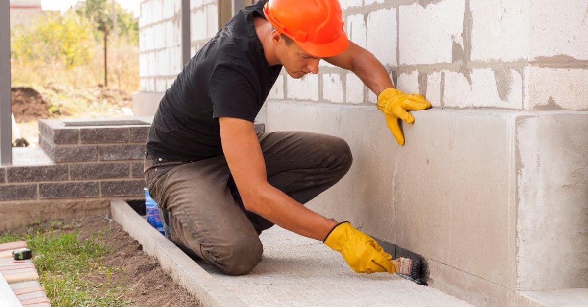 A man wearing an orange hard hat and gloves brushing a black coating onto the base of a home's foundation.