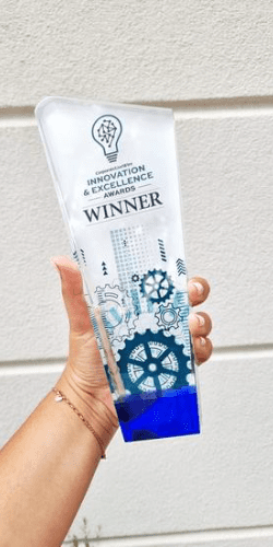 Photo of a hand holding a tall, transparent award with blue detailing at the bottom. The award features a lightbulb icon and is labeled 'Innovation & Excellence Awards' with the word 'WINNER' prominently displayed. The design also includes gear and circuit patterns, symbolizing innovation and technology